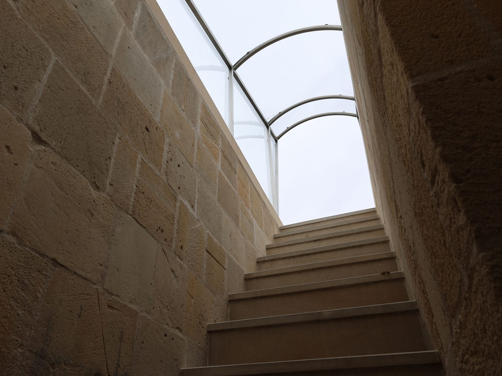 A stone staircase rising between old masonry walls toward a glazed roof canopy.