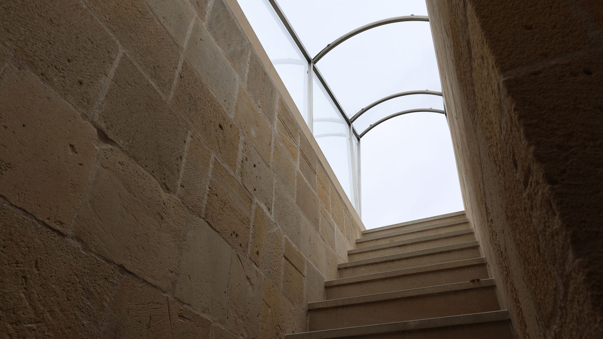 A stone staircase rising between old masonry walls toward a glazed roof canopy.